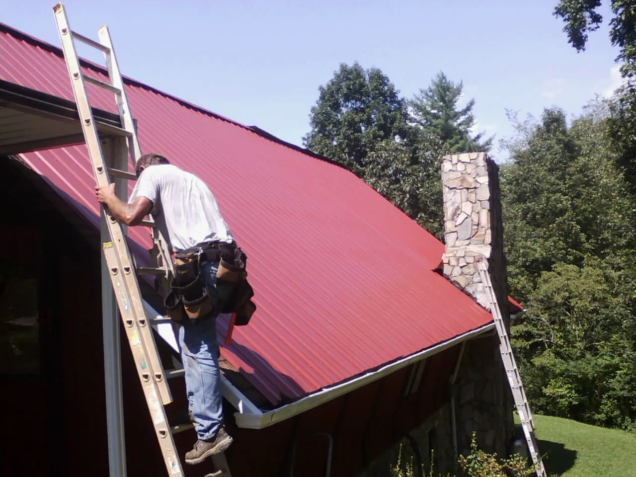 Roofer on a ladder beside a red metal roof and stone chimney — Soffit & Fascia Repair by Roofing Brothers in Campobello